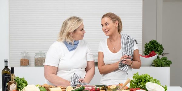 Two women in a kitchen preparing fresh vegetables and cheese with olive oil and spices.