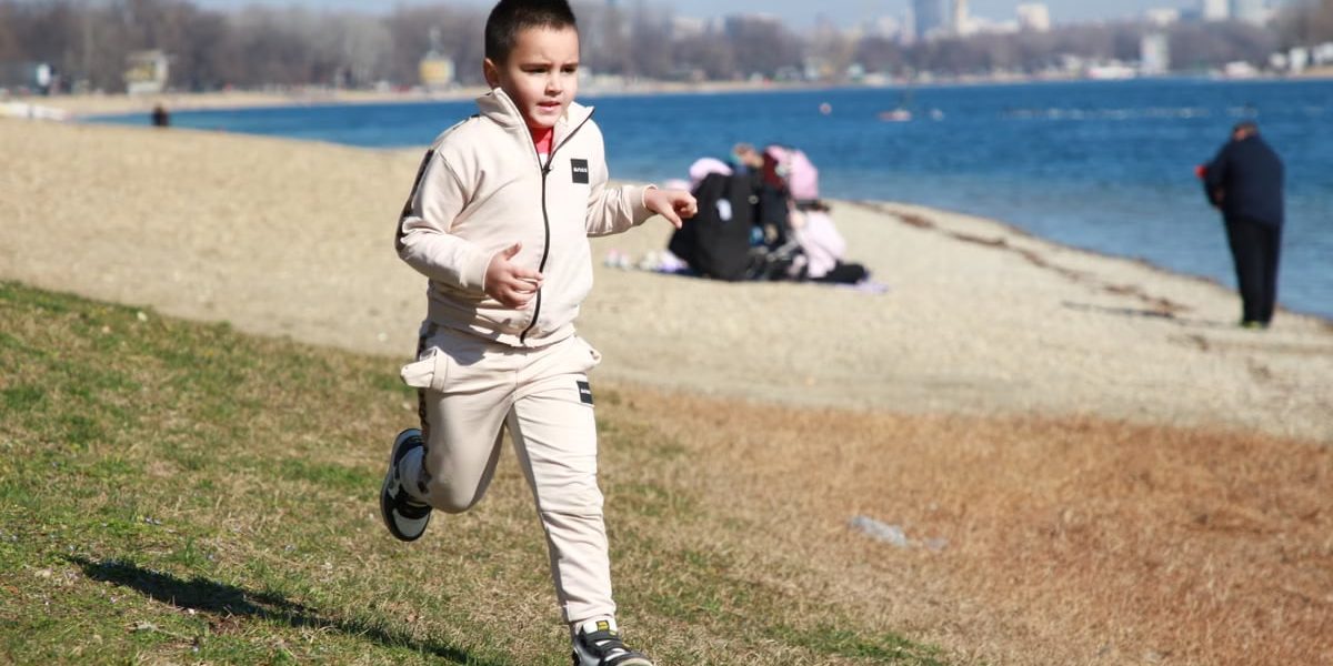 Young boy in a tracksuit runs energetically by the waterfront, embodying the joy of a sunny day outdoors.