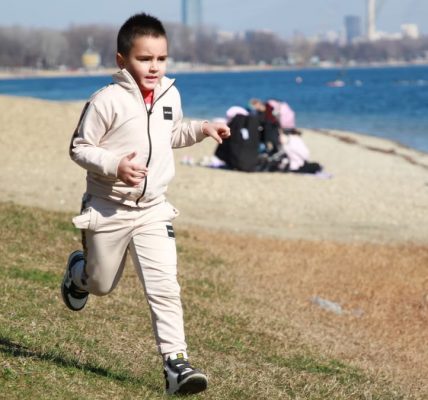 Young boy in a tracksuit runs energetically by the waterfront, embodying the joy of a sunny day outdoors.