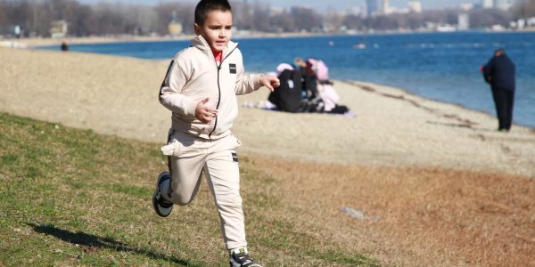 Young boy in a tracksuit runs energetically by the waterfront, embodying the joy of a sunny day outdoors.